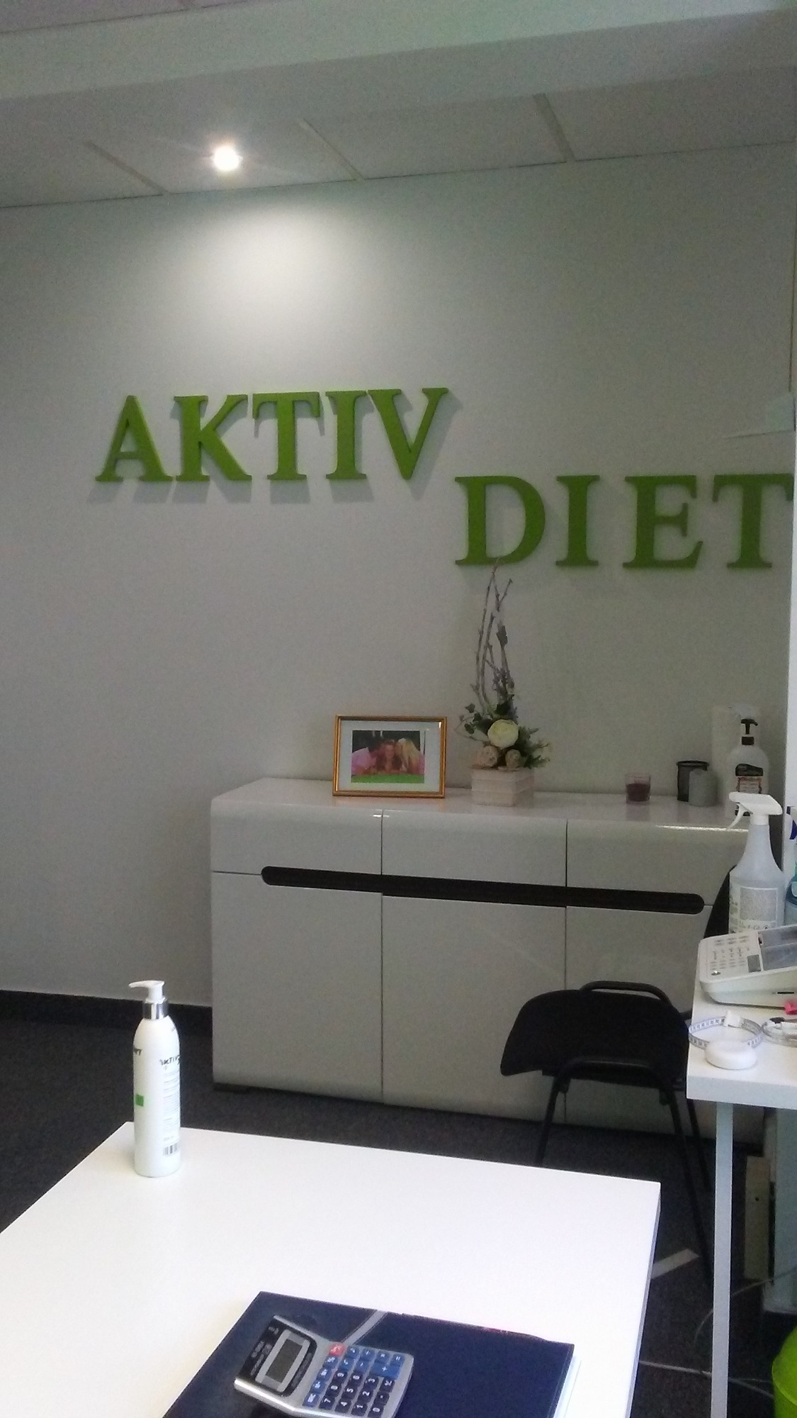 Wnętrze gabinetu z białymi meblami, dekoracjami kwiatowymi i napisem 'AKTIV DIET' na ścianie. Na pierwszym planie biały stół z kalkulatorem i butelką płynu.