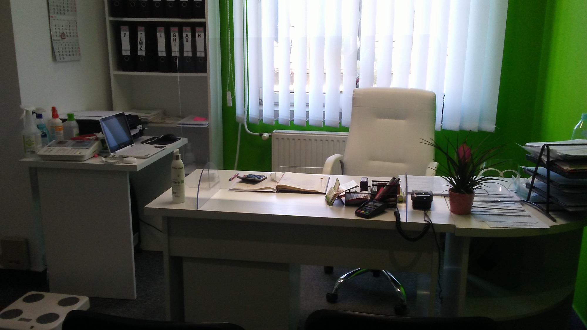 Jasne biuro z białym biurkiem, na którym leżą kalkulator, otwarty notatnik, telefon, organizer na długopisy i roślina doniczkowa. W tle białe krzesło, okno z żaluzjami i regał z segregatorami.