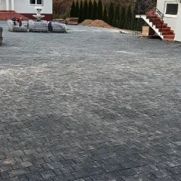 Ułożona szara kostka brukowa na podjeździe, widoczne palety z kostką, biały dom z czerwonymi schodami i ozdobną fontanną w tle.