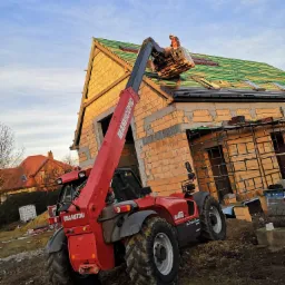 Czerwony podnośnik teleskopowy Manitou z operatorem na platformie, układający dachówkę na budynku w stanie surowym otwartym, widok z dołu na tle błękitnego nieba.