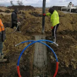 Wylewanie betonu do wąskiego wykopu pod fundament z widocznym zbrojeniem i rurami instalacyjnymi w kolorze niebiesko-czerwonym, pracownicy w odzieży roboczej, betonomieszarka w tle.