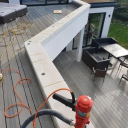 Czerwona wiertnica rdzeniowa na murku z jasnych bloczków z wywierconym otworem, na tle tarasu z desek kompozytowych i nowoczesnego domu z zielonym dachem.