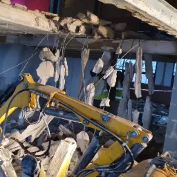 Żółta maszyna budowlana kruszy betonową konstrukcję, z wiszącymi fragmentami gruzu, w tle widoczna postać w kamizelce odblaskowej.