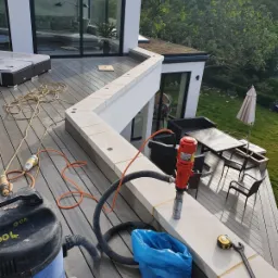 Wiercenie otworów w kamiennej balustradzie tarasu nowoczesnego domu z użyciem wiertnicy diamentowej, widoczny odkurzacz przemysłowy, narzędzia pomiarowe i worek na odpady.