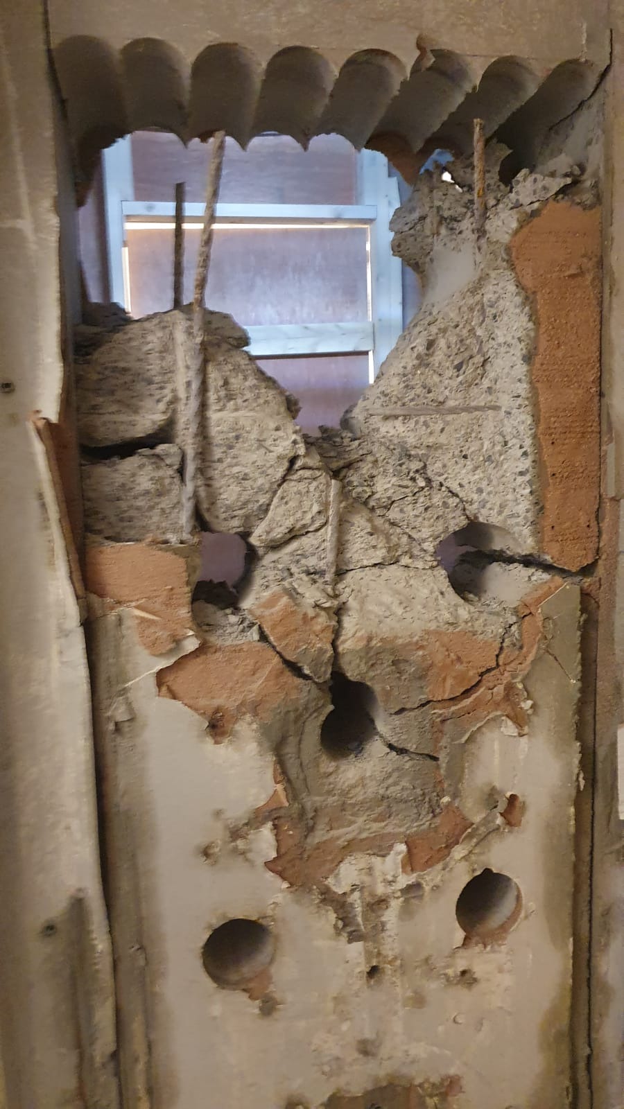 Fragment zdemontowanej ściany z widocznymi otworami, cegłami, betonem i wystającymi prętami zbrojeniowymi w otworze okiennym.