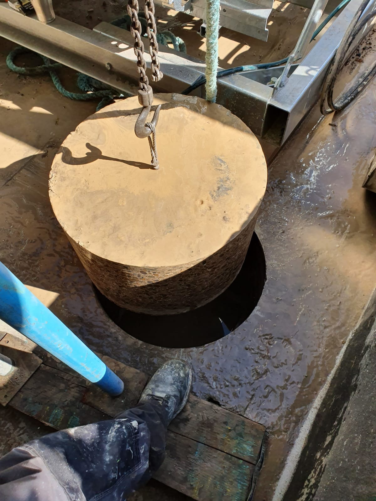 Ciężki, okrągły, betonowy element zawieszony na łańcuchu nad otworem w mokrej, betonowej powierzchni; widoczny fragment nogi pracownika w ubrudzonym bucie.