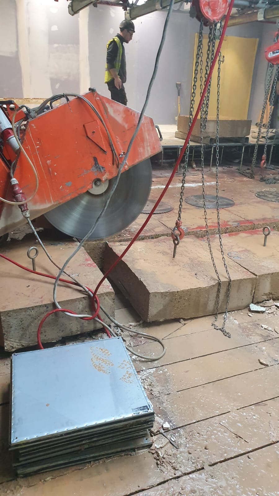 Pomarańczowa piła do betonu tnie gruby betonowy blok na brudnej podłodze, z robotnikiem w tle i łańcuchami podtrzymującymi element.