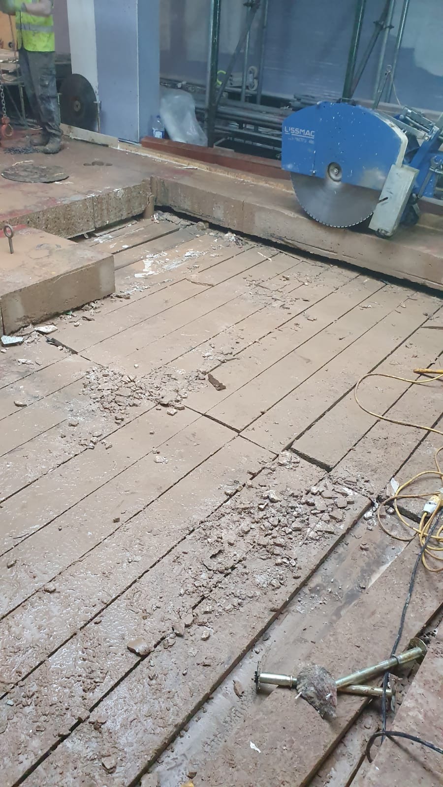 Cięcie betonowej podłogi za pomocą piły Lissmac, widoczne fragmenty gruzu i pracownik w odblaskowej kamizelce w tle.