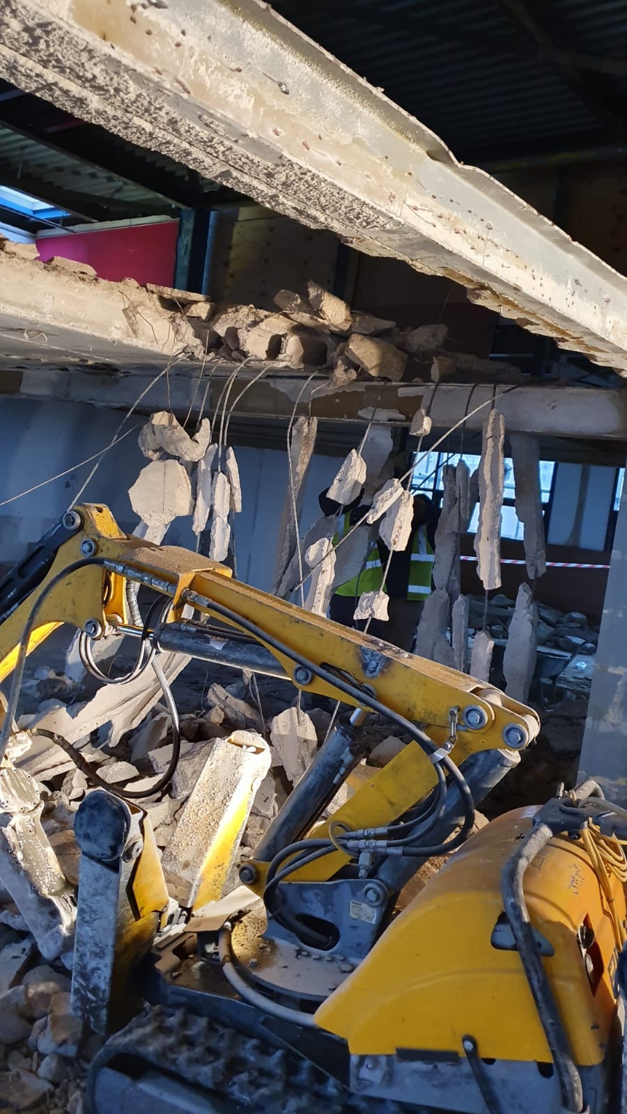Żółta maszyna budowlana kruszy betonową konstrukcję, z wiszącymi fragmentami gruzu, w tle widoczna postać w kamizelce odblaskowej.
