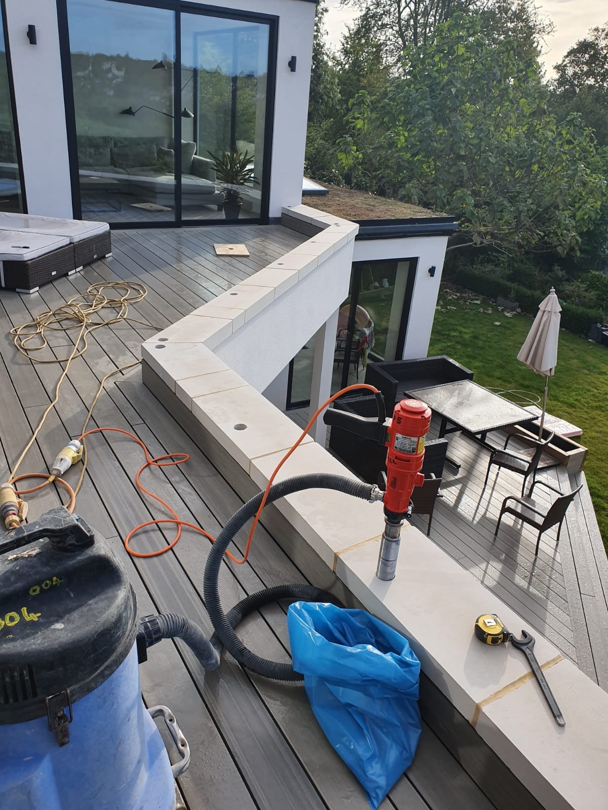 Wiercenie otworów w kamiennej balustradzie tarasu nowoczesnego domu z użyciem wiertnicy diamentowej, widoczny odkurzacz przemysłowy, narzędzia pomiarowe i worek na odpady.