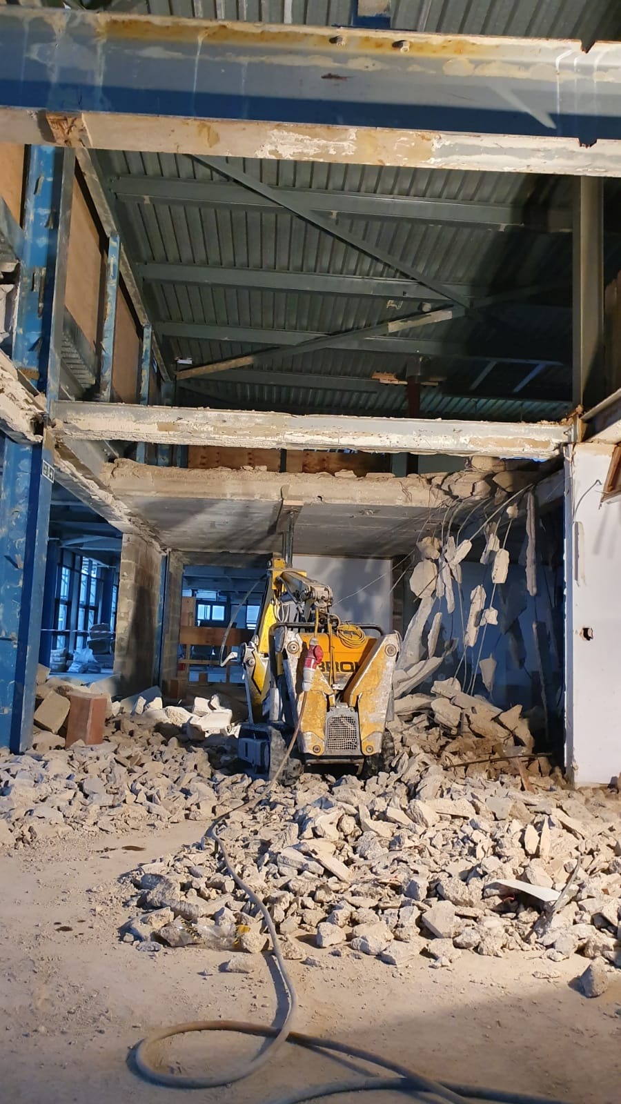 Żółta maszyna do prac rozbiórkowych w trakcie usuwania fragmentu ściany wewnątrz budynku, gruz na podłodze, widoczne elementy konstrukcyjne budynku.