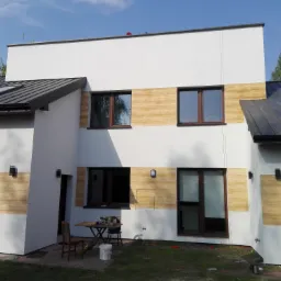 Nowoczesny dom z białymi ścianami i panelami imitującymi drewno, czarnym dachem z oknami dachowymi, widoczny stół z krzesłem i wiadro farby na trawniku.