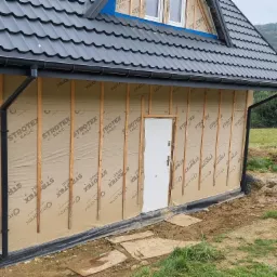 Dach z blachy trapezowej w kolorze grafitowym i system rynnowy zamontowany na domu szkieletowym w trakcie budowy, widoczne ściany osłonowe z płyt OSB i izolacji.