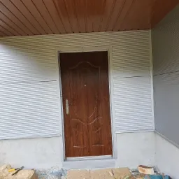 Wejście do budynku z brązowymi drzwiami i srebrnymi ścianami z paneli, widoczne narzędzia murarskie i fragment krajobrazu