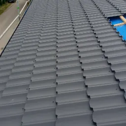 Szary dach z blachodachówki modułowej, widoczne łączenia i wkręty, w tle rusztowanie i zieleń drzew.