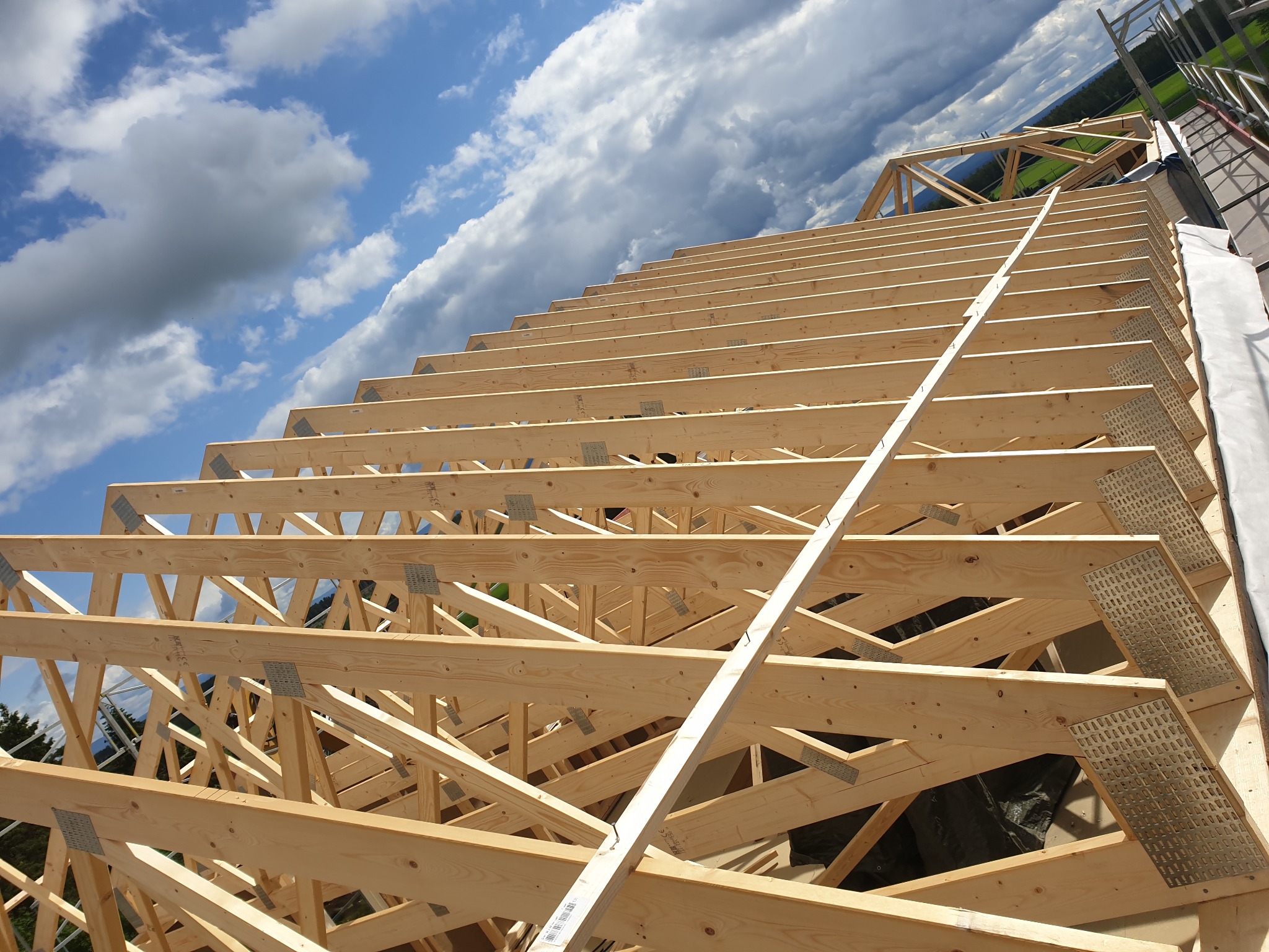 Konstrukcja drewniana więźby dachowej z widocznymi elementami łączącymi, na tle błękitnego nieba z obłokami.