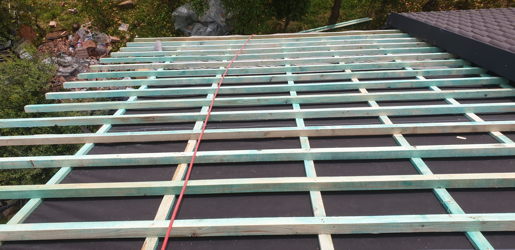 Konstrukcja drewniana dachu pokryta czarną membraną, widoczne łatki pomalowane na niebiesko-zielony kolor, w tle gruz i pozostałości po starej dachówce.