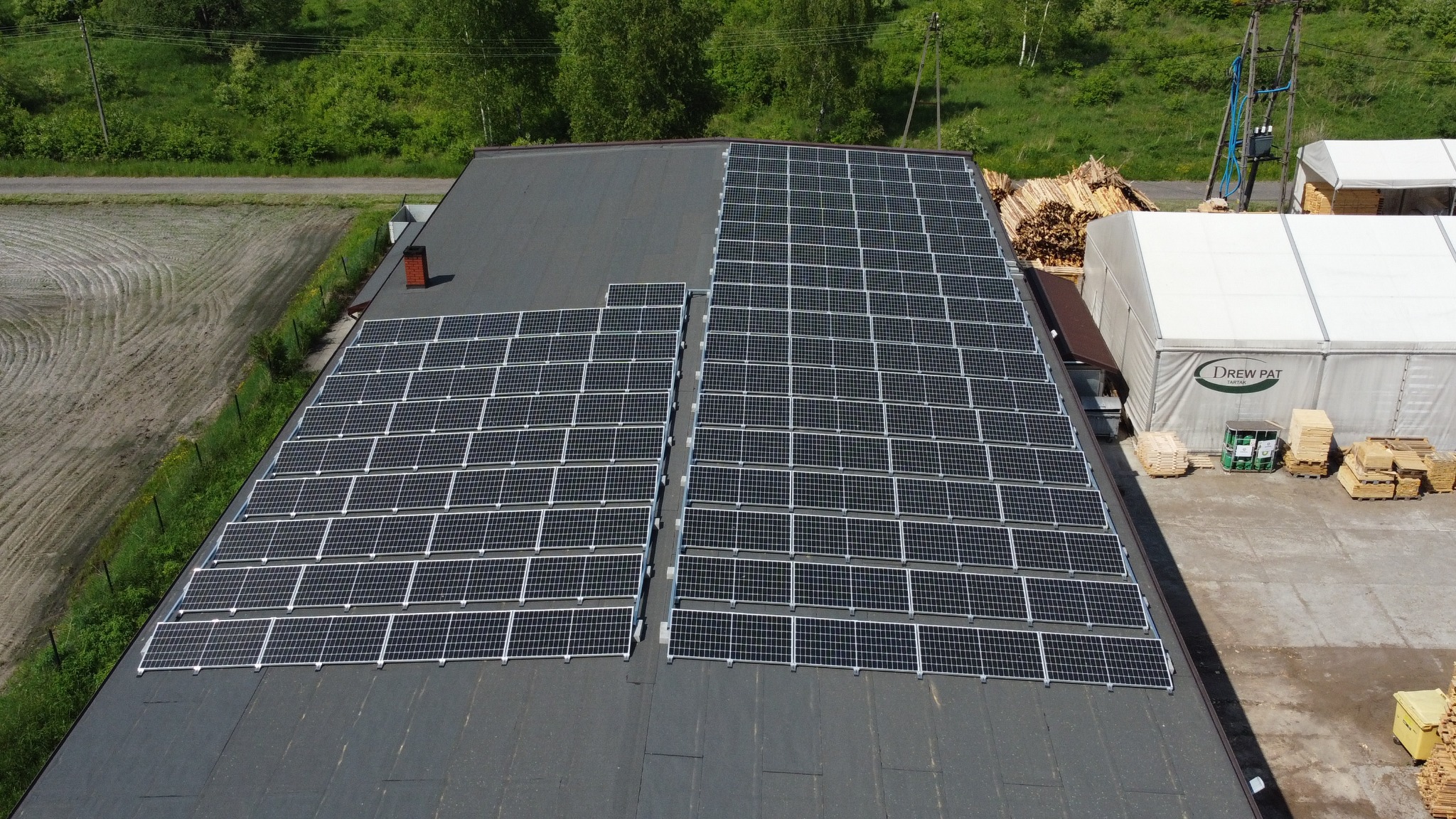 Widok z góry na dużą instalację fotowoltaiczną na dachu przemysłowym. Panele ułożone rzędami, obok składowane drewno i namiot magazynowy. Otoczenie zielone, drzewa w tle.