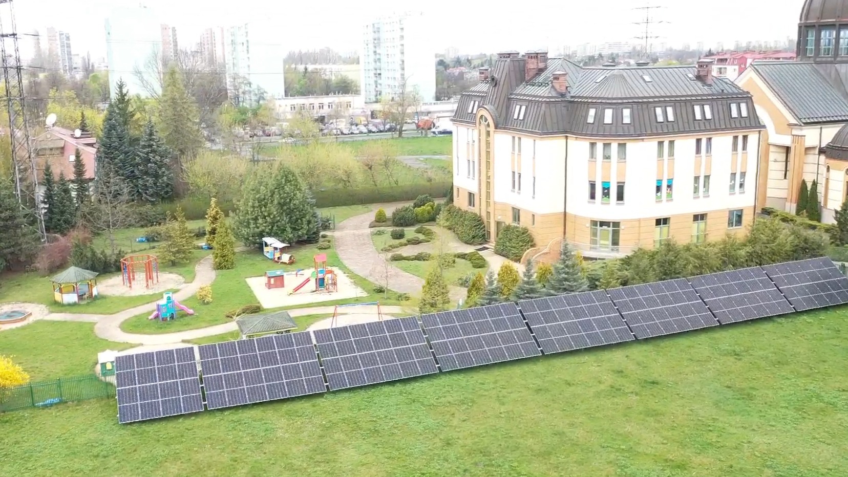Instalacja fotowoltaiczna na trawniku przed budynkiem z placem zabaw. Panele ułożone rzędami, widok z góry, budynek w tle, zielony trawnik.