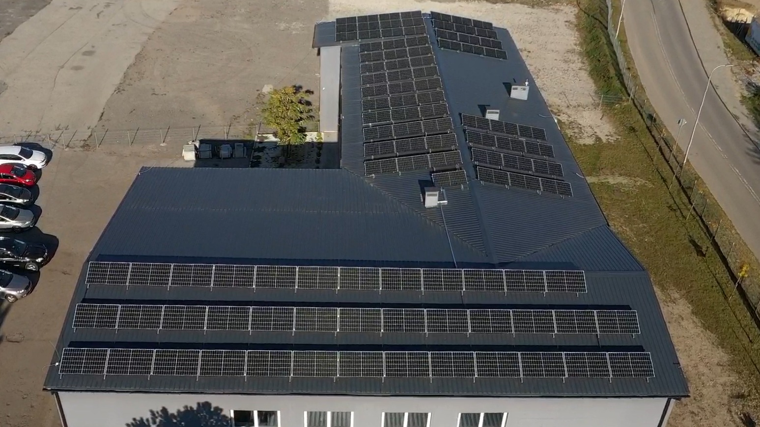 Widok z drona na budynek z panelami słonecznymi na dachu w Katowicach. Rzędy paneli ułożone na różnych poziomach dachu. Obok budynku zaparkowane samochody.