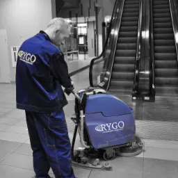 Pracownik w niebieskim uniformie z logo firmy RYGO czyści podłogę w Baranowie za pomocą profesjonalnej maszyny czyszczącej w pobliżu ruchomych schodów w budynku.