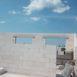 Wznoszenie ścian domu z bloczków z betonu komórkowego, widoczne otwory okienne z nadprożami, murarz przy pracy, błękitne niebo w tle.