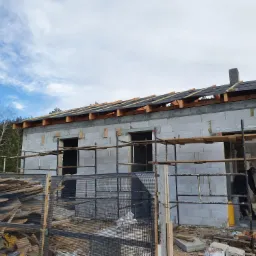 Surowy stan budynku z białych bloczk&oacute;w, widoczne rusztowania, przygotowane drewno na dachu i stos desek na pierwszym planie, częściowo zasłonięty siatką.