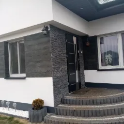 Wykończona elewacja domu jednorodzinnego z elementami imitującymi drewno i kamień, widoczne schody wejściowe i oświetlenie zewnętrzne.