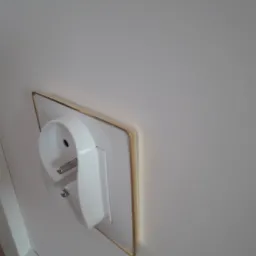 Białe gniazdko elektryczne z cienką złotą ramką na białej ścianie z białą listwą przypodłogową, widok z boku.