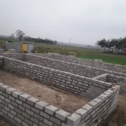 Wznoszone ściany fundamentowe z bloczków betonowych na działce budowlanej w pochmurny dzień, widoczne elementy szalunku i zaplecze budowy w tle.