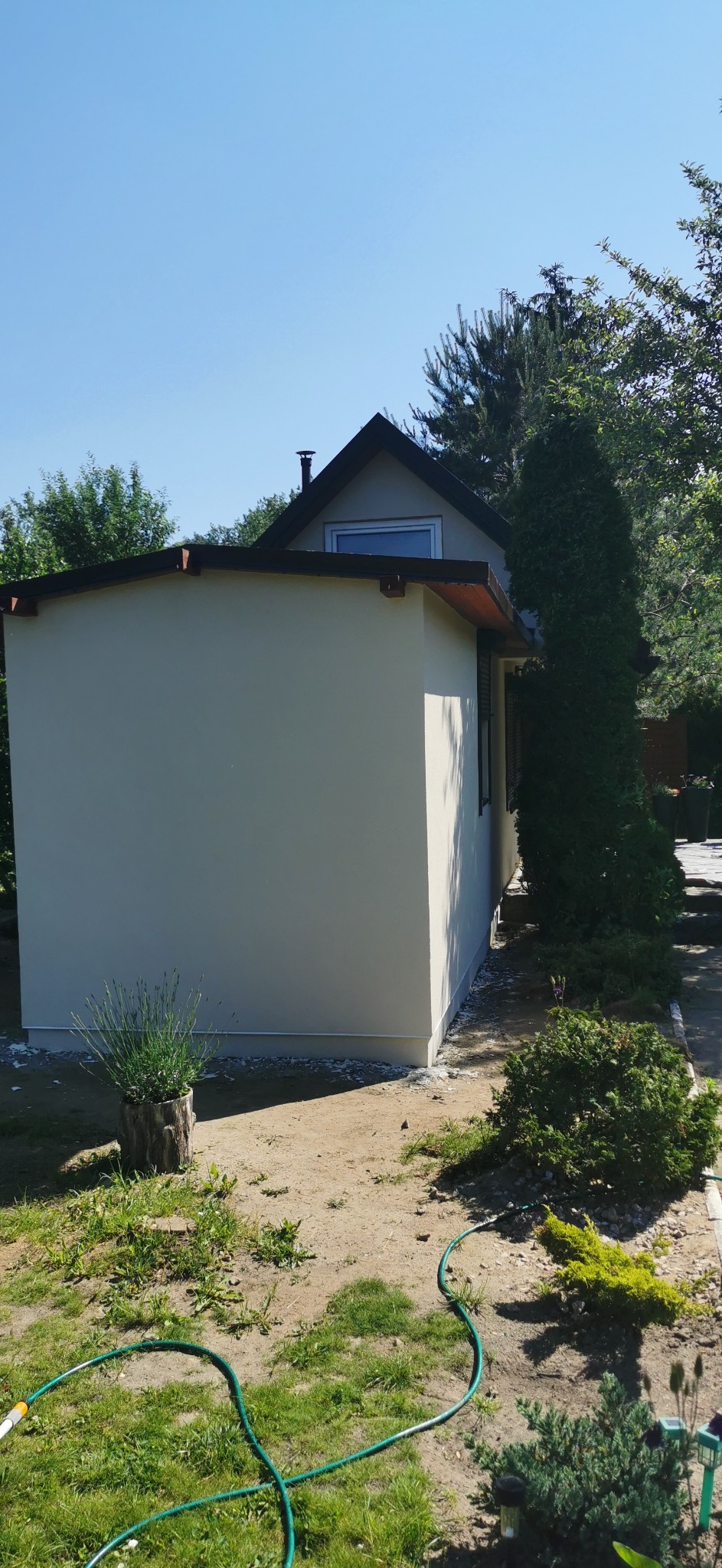 Świeżo otynkowana elewacja budynku z oknem na poddaszu, widoczna z boku, z zielonym wężem ogrodowym leżącym na trawie w pobliżu.