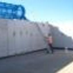 Phu.Jon-Bud Joanna Jagielska - Pracownik w pomarańczowej kamizelce opiera się o wysoki, szary fundament budynku w trakcie budowy, widoczne ślady po szalunkach.