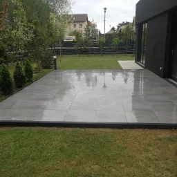 Nowoczesny taras z szarych płyt, po deszczu, przylegający do ciemnej bryły domu, z widokiem na zadbany trawnik i ogrodzenie.