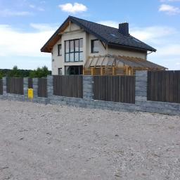 Phu.Jon-Bud Joanna Jagielska - Nowoczesne ogrodzenie panelowe z elementami betonowymi na tle domu jednorodzinnego z szarą dachówką i drewnianą konstrukcją z tyłu, widoczne niebo z chmurami.