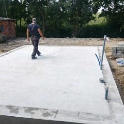 Phu.Jon-Bud Joanna Jagielska - Wylana płyta fundamentowa z widocznymi wystającymi rurami instalacyjnymi i pracownikiem w ubraniu roboczym, w tle budynek gospodarczy z czerwonej cegły.