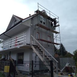 Phu.Jon-Bud Joanna Jagielska - Dwukondygnacyjny dom w trakcie ocieplania, z szarym tynkiem i białym balkonem, otoczony rusztowaniem, na którym pracują dwie osoby, widoczny zza czarnego metalowego ogrodzenia.