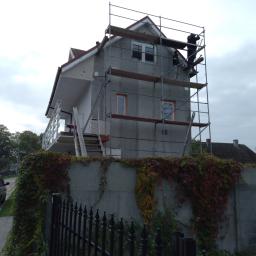 Phu.Jon-Bud Joanna Jagielska - Dwupiętrowy dom w trakcie ocieplania, otoczony rusztowaniem, z widocznymi pracownikami na górnej kondygnacji, ogrodzony betonowym murem porośniętym winobluszczem i metalowym płotem.