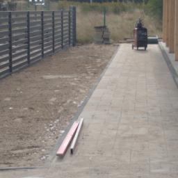 Phu.Jon-Bud Joanna Jagielska - Szara, drewniana palisada ogrodzeniowa, świeżo ułożona kostka brukowa na ścieżce, poziomice na kostce, w tle spawarka i drewniane słupy konstrukcyjne.