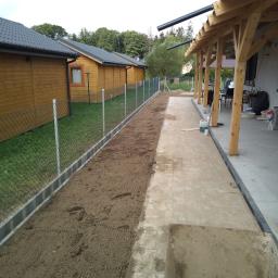 Phu.Jon-Bud Joanna Jagielska - Przygotowanie terenu pod chodnik z kostki brukowej i montaż ogrodzenia siatkowego przy drewnianych domkach letniskowych, widoczne krawężniki i wyrównana powierzchnia gruntu.