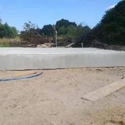 Wylany betonowy fundament płyty pod budynek jednorodzinny, widoczny niebieski wąż doprowadzający wodę, teren budowy w otoczeniu zieleni.