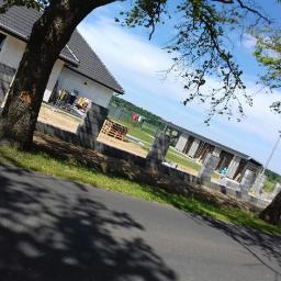 Phu.Jon-Bud Joanna Jagielska - Budowa ogrodzenia z betonowych bloczków wokół nowo wybudowanego domu jednorodzinnego, widok z drogi, częściowo zasłonięty przez drzewa, w tle widoczny teren zielony.