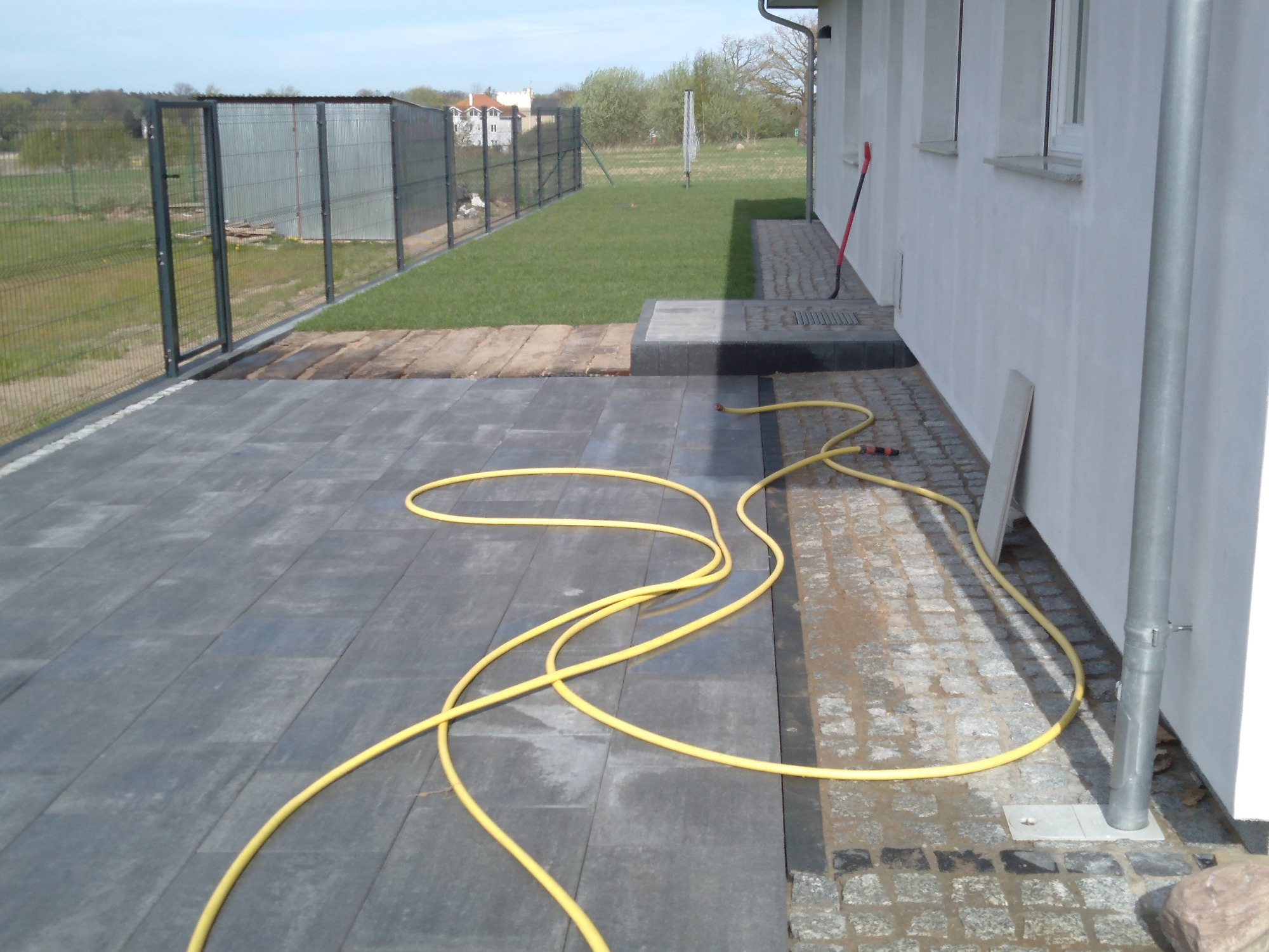 Nowoczesny taras z szarych płyt betonowych, obrzeże z kostki granitowej, żółty wąż ogrodowy na powierzchni, widok na ogrodzenie panelowe i trawnik przy domu.