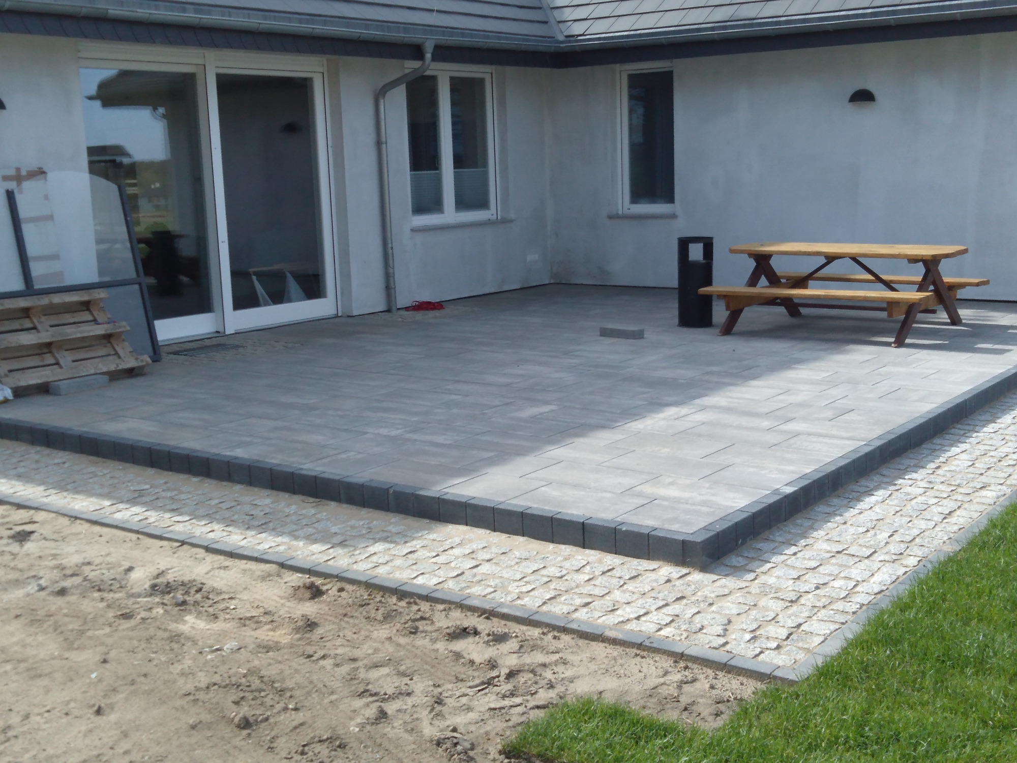Nowoczesny taras z szarych płyt betonowych obramowany ciemniejszą kostką, przechodzący w ścieżkę z jasnej kostki granitowej, z drewnianym stołem piknikowym w tle i elementami domu.