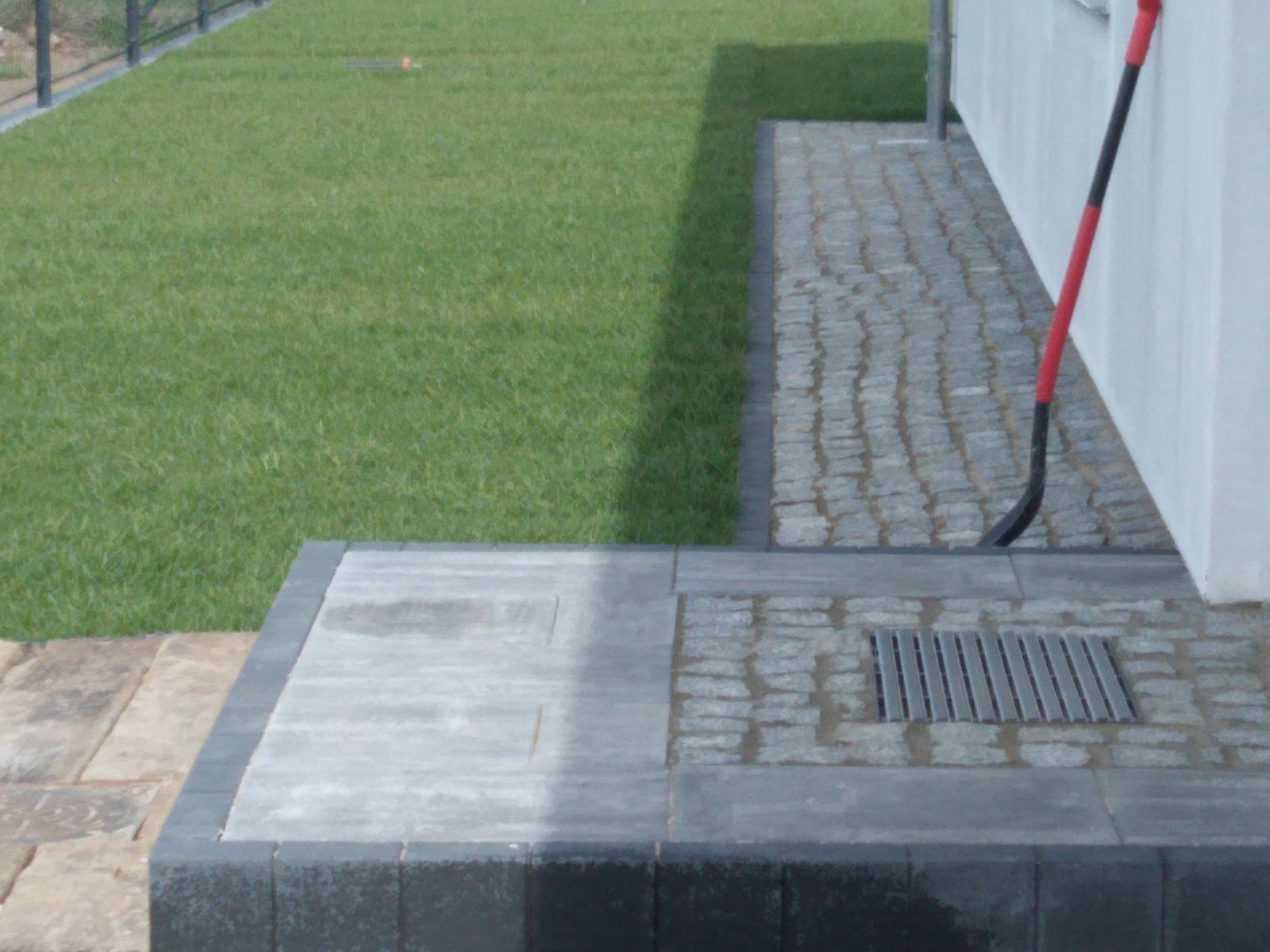 Świeżo ułożona kostka brukowa w odcieniach szarości z odpływem liniowym, sąsiadująca z trawnikiem i kostką granitową, z łopatą opartą o ścianę budynku.