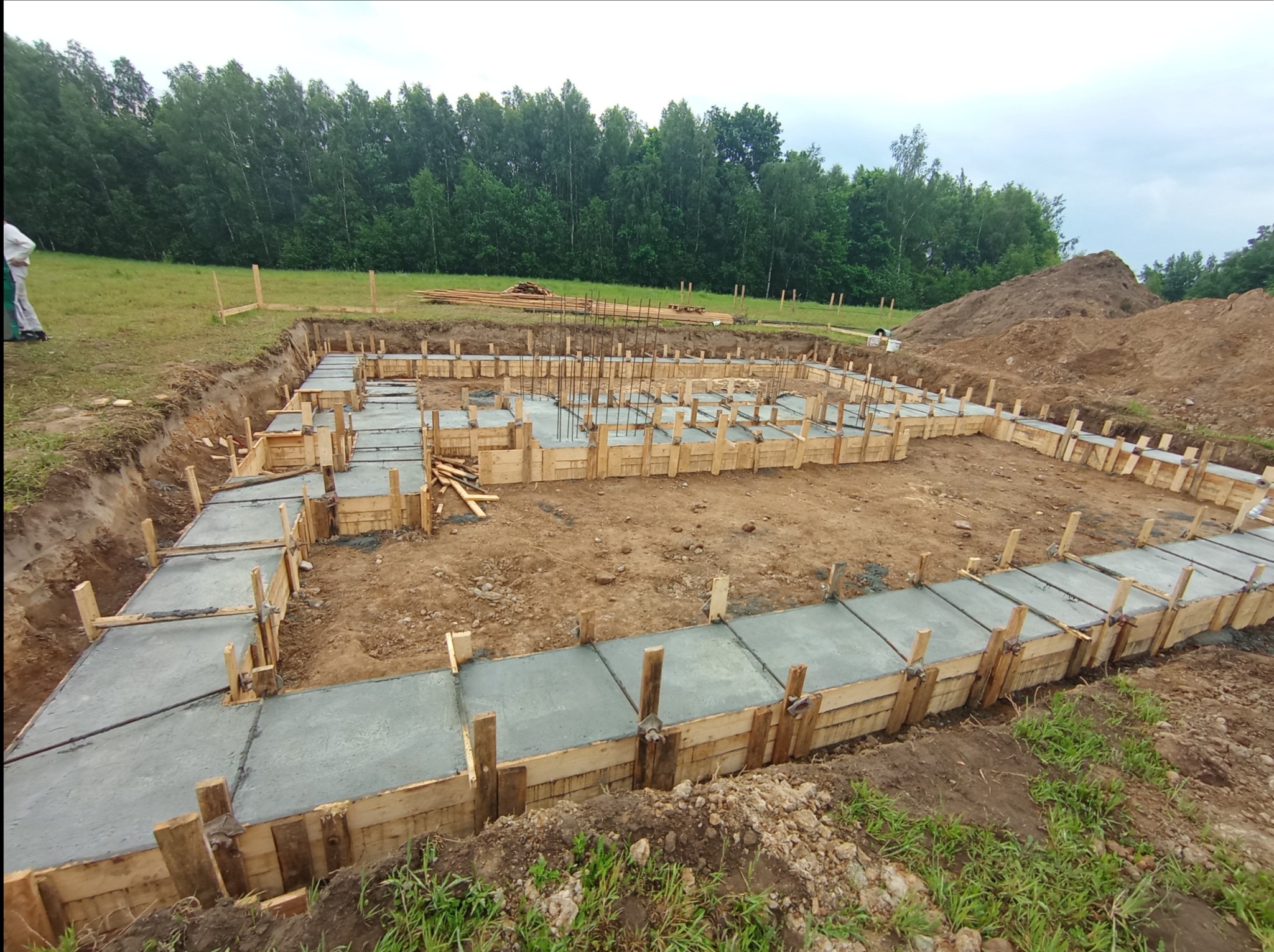 Wylane fundamenty pod budynek mieszkalny z drewnianym szalunkiem, widoczne zbrojenia i wykopana ziemia, w tle las.