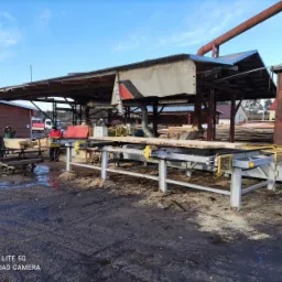 Praca w toku: deski transportowane na automatycznej linii produkcyjnej pod zadaszeniem, w tle wózek widłowy i pracownicy w ubraniach roboczych.