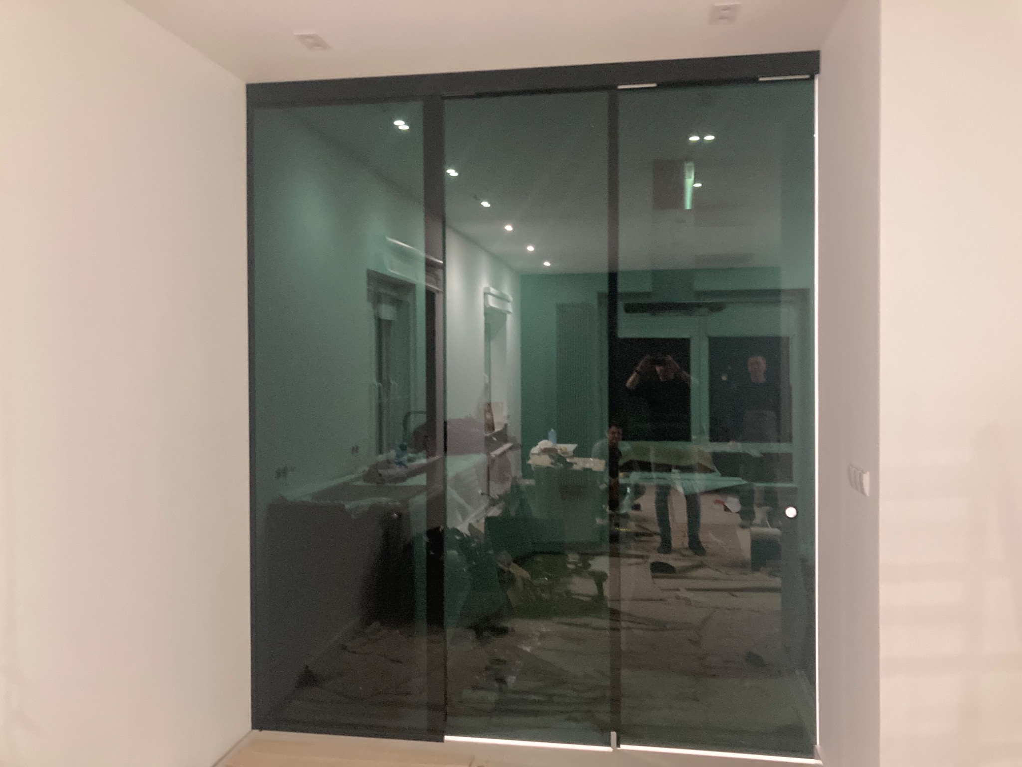 Szklane drzwi przesuwne w czarnej ramie, widoczny remont w tle i odbicie fotografa.