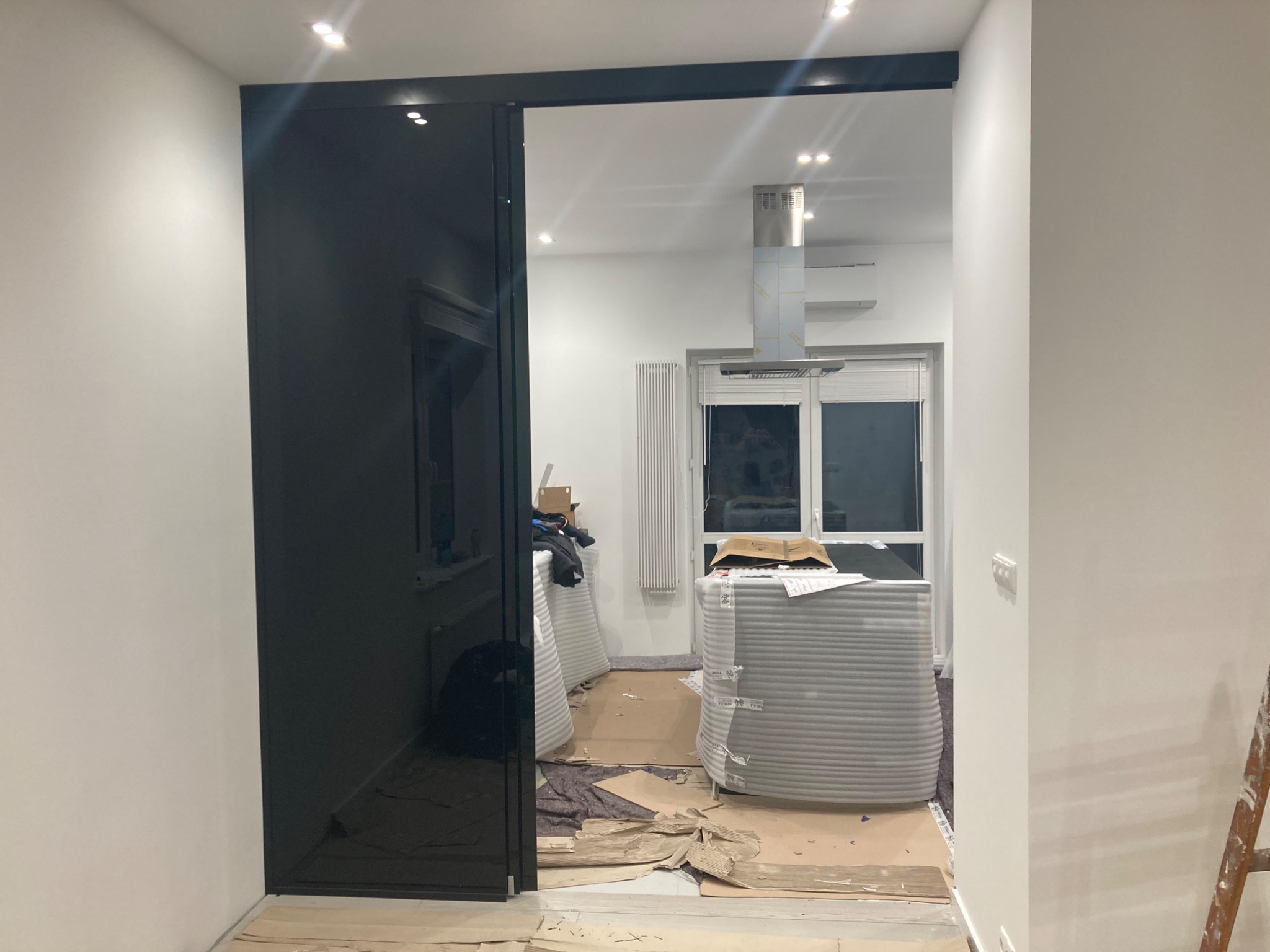 Czarne, szklane drzwi przesuwne z widocznym wnętrzem w trakcie remontu: zabezpieczone meble, kartony na podłodze, drabina oparta o ścianę.