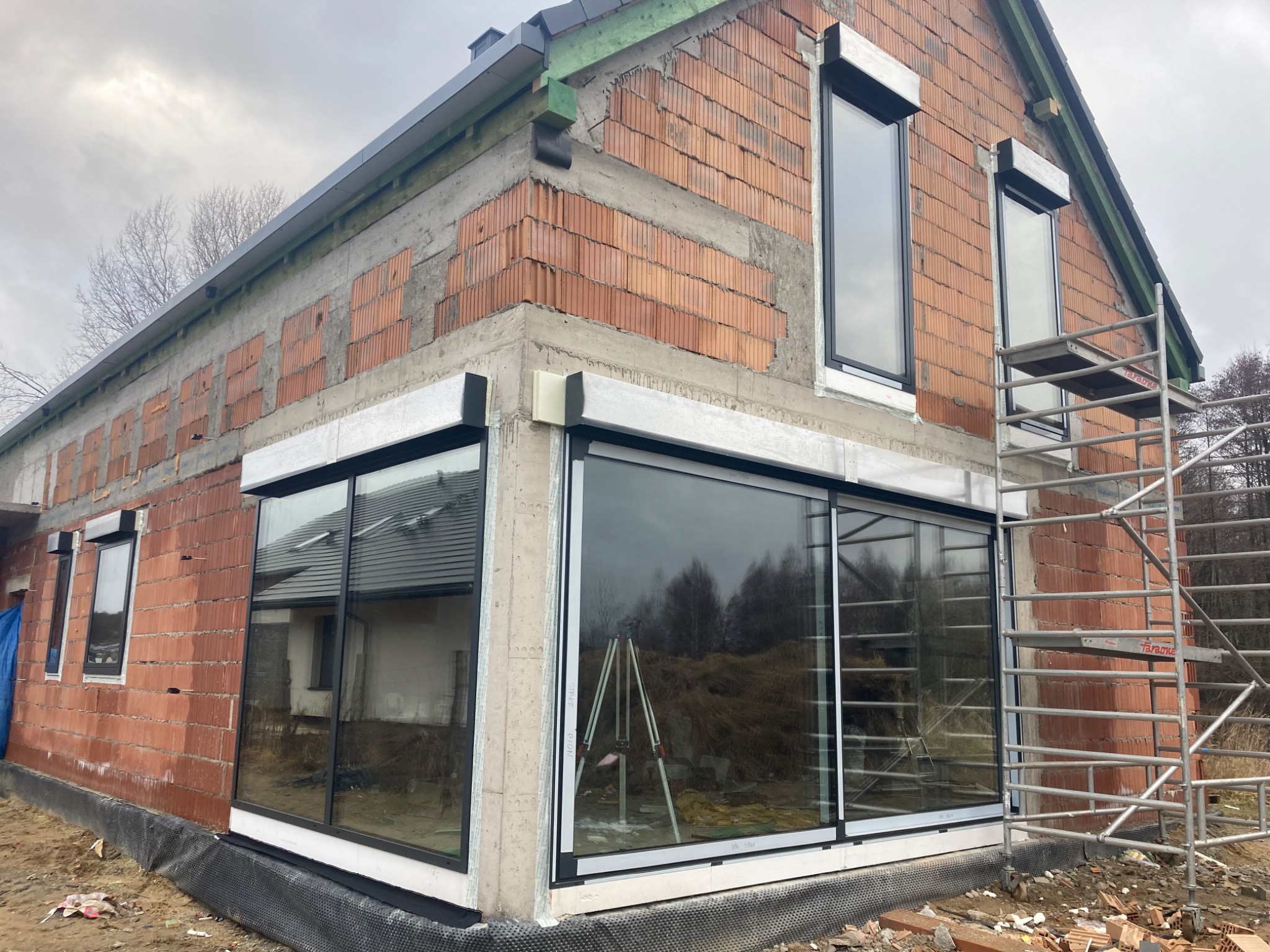 Dom w trakcie budowy z zamontowanymi dużymi przesuwnymi oknami, widoczne rusztowanie i niewykończona elewacja z cegły.