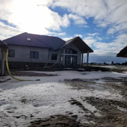 Surowy budynek jednorodzinny w trakcie budowy, z szarymi tynkami zewnętrznymi i ciemnym dachem na tle pochmurnego nieba, widoczne żółte węże na zewnątrz budynku, resztki śniegu na ziemi.